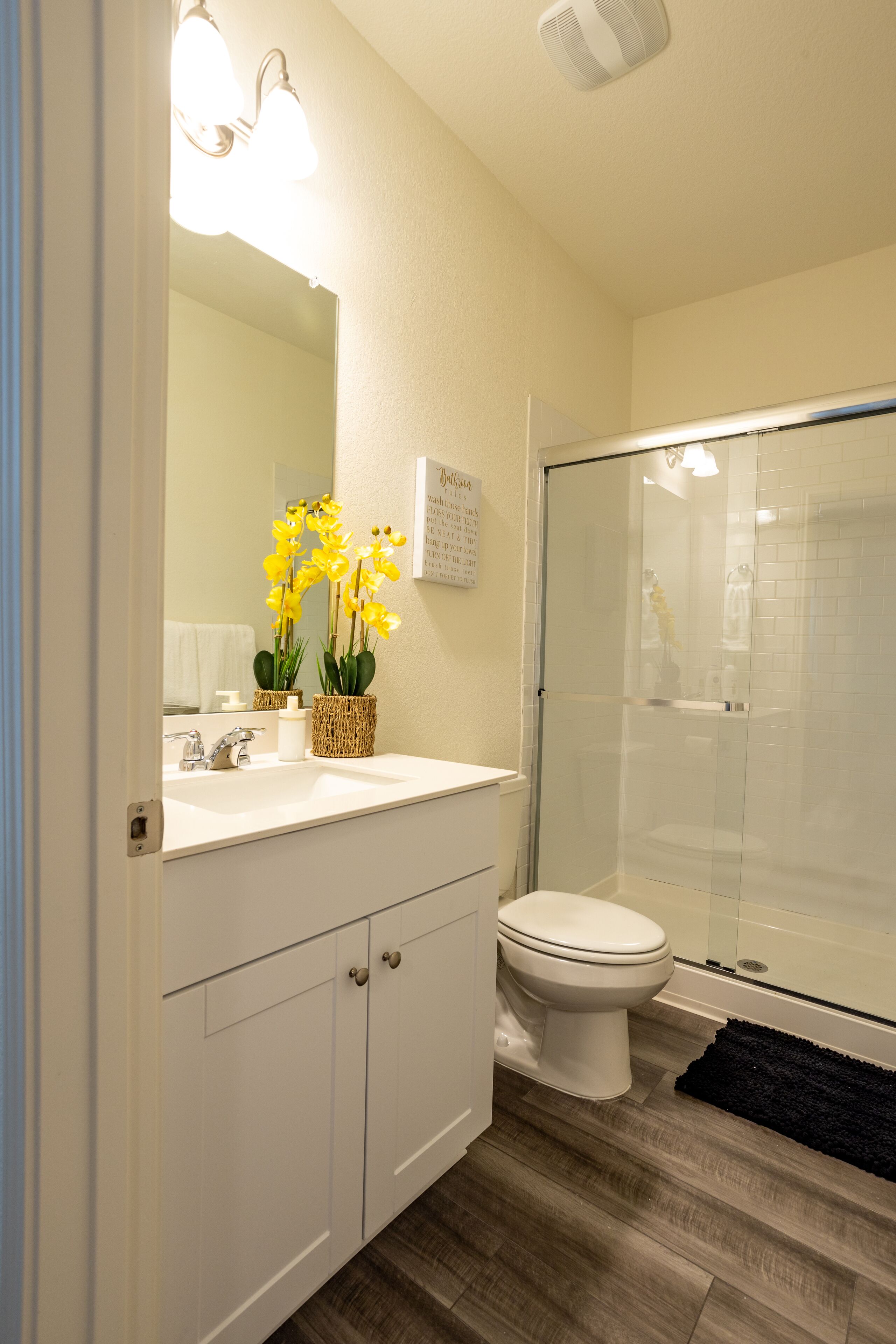 A bathroom with a white sink, a toilet, a glass shower enclosure, and a dark rug on the floor.