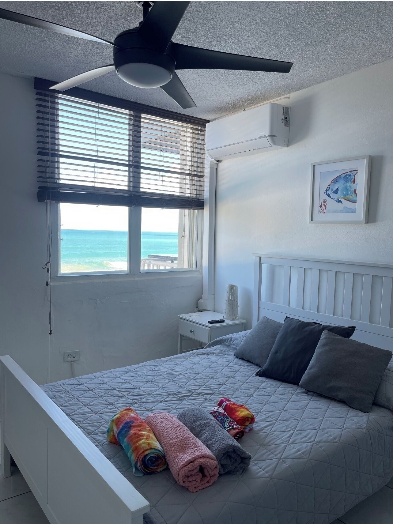 Une chambre à coucher avec un lit, un ventilateur de plafond, une fenêtre avec des stores, une table de chevet et un tableau encadré au mur.