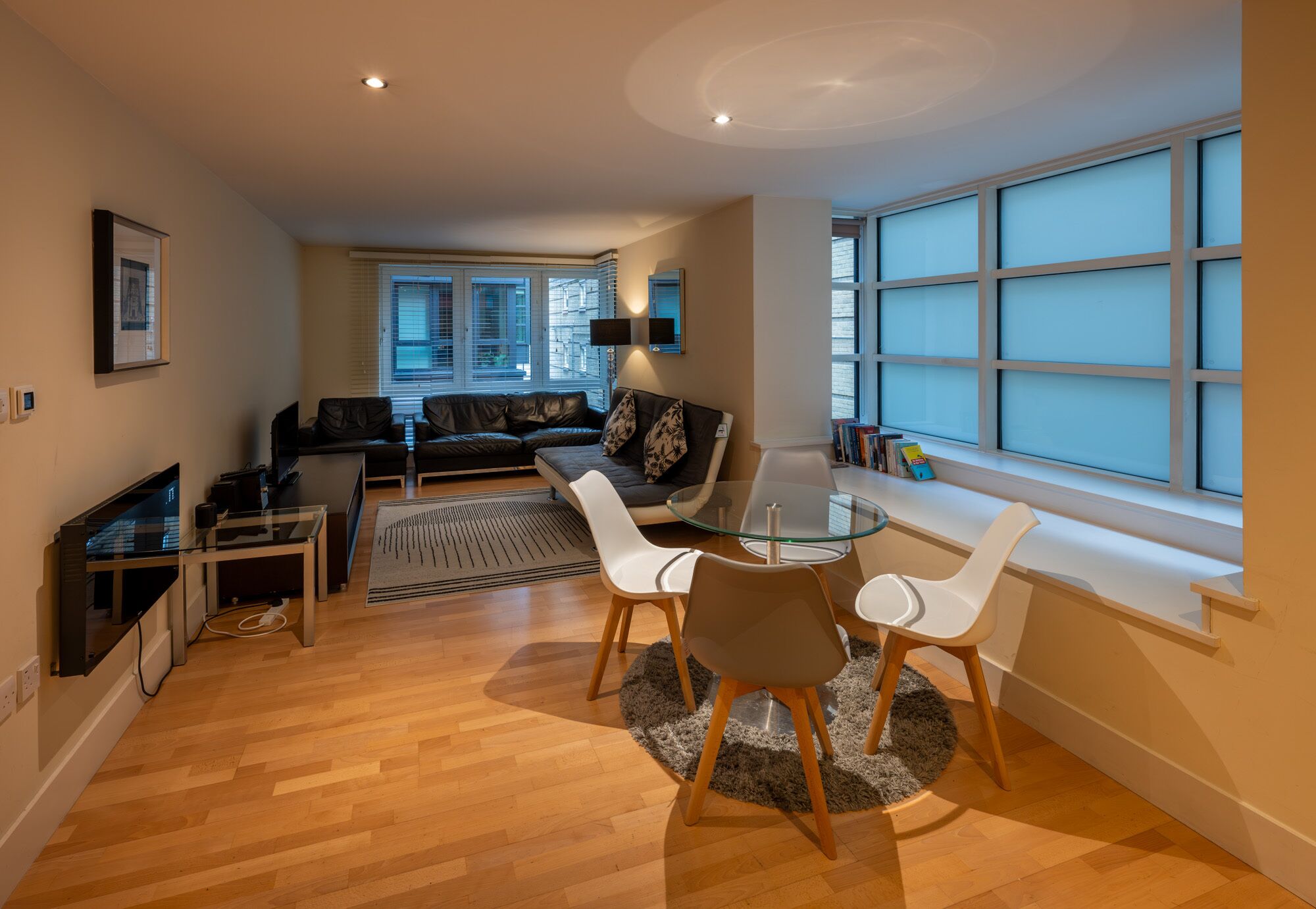 A modern living room with a glass dining table, white chairs, a leather sofa, and a large window with blinds.