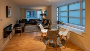 Un salon moderne avec une table à manger en verre, des chaises blanches, un canapé en cuir et une grande fenêtre avec des stores.