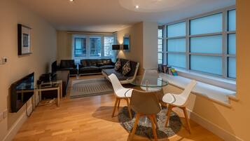 A modern living room with a glass dining table, white chairs, a leather sofa, and a large window with blinds.