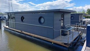 Een stilstaande woonboot met een vlak dek, twee ronde ramen en een naambordje met de tekst 'LA MARE'.