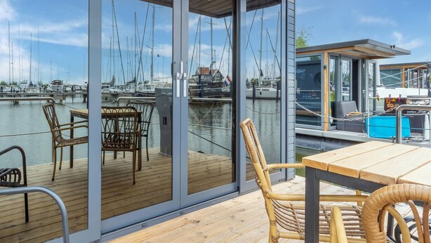 Een modern terras buiten met glazen deuren, rieten meubels en uitzicht op een jachthaven met boten.