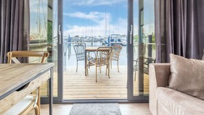 Een kamer met een houten tafel en stoelen, een bank en een schuifdeur naar een balkon met uitzicht op een jachthaven.