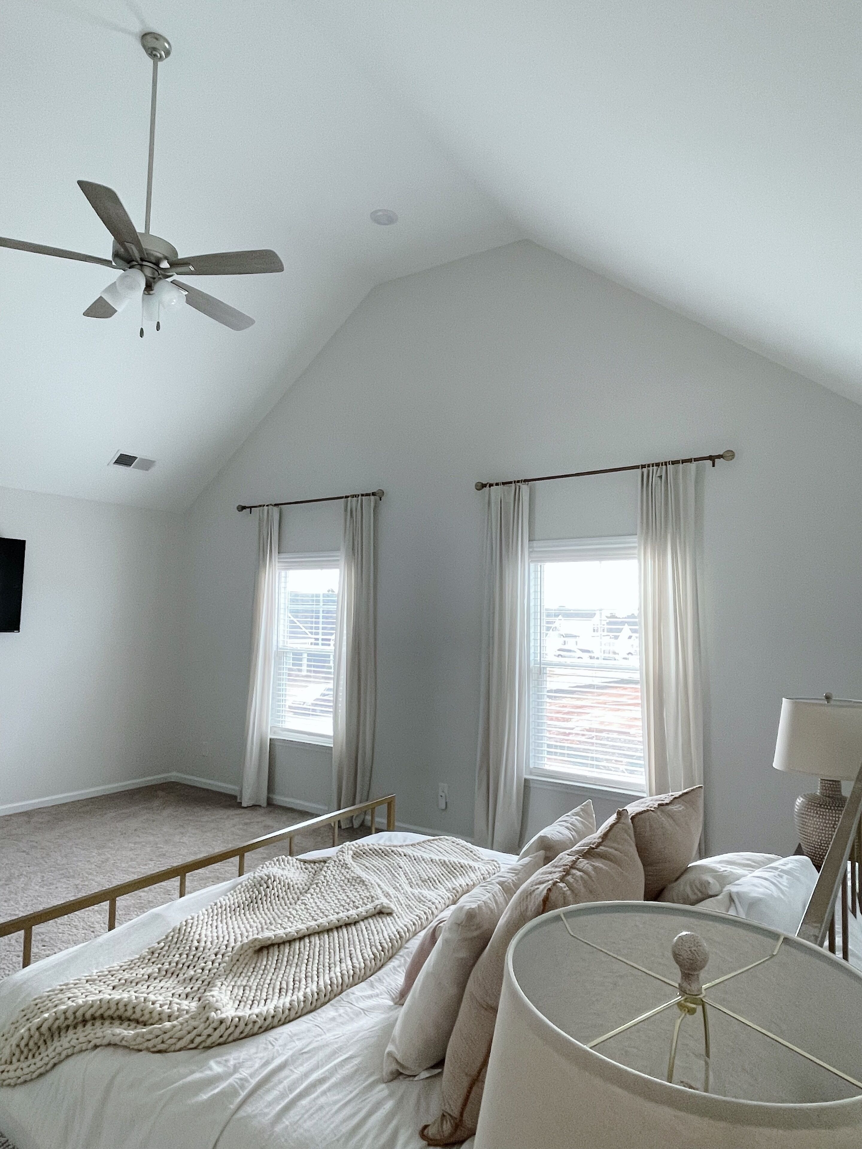 Une chambre avec un plafond en pente, deux fenêtres avec des rideaux, un lit recouvert d’une couverture tricotée et un ventilateur de plafond.