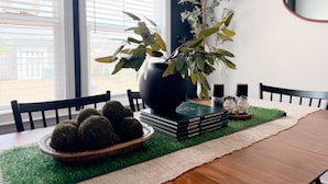Une table à manger avec un set de table vert, un bol rempli d’objets ronds, un vase avec des branches et une pile de livres.