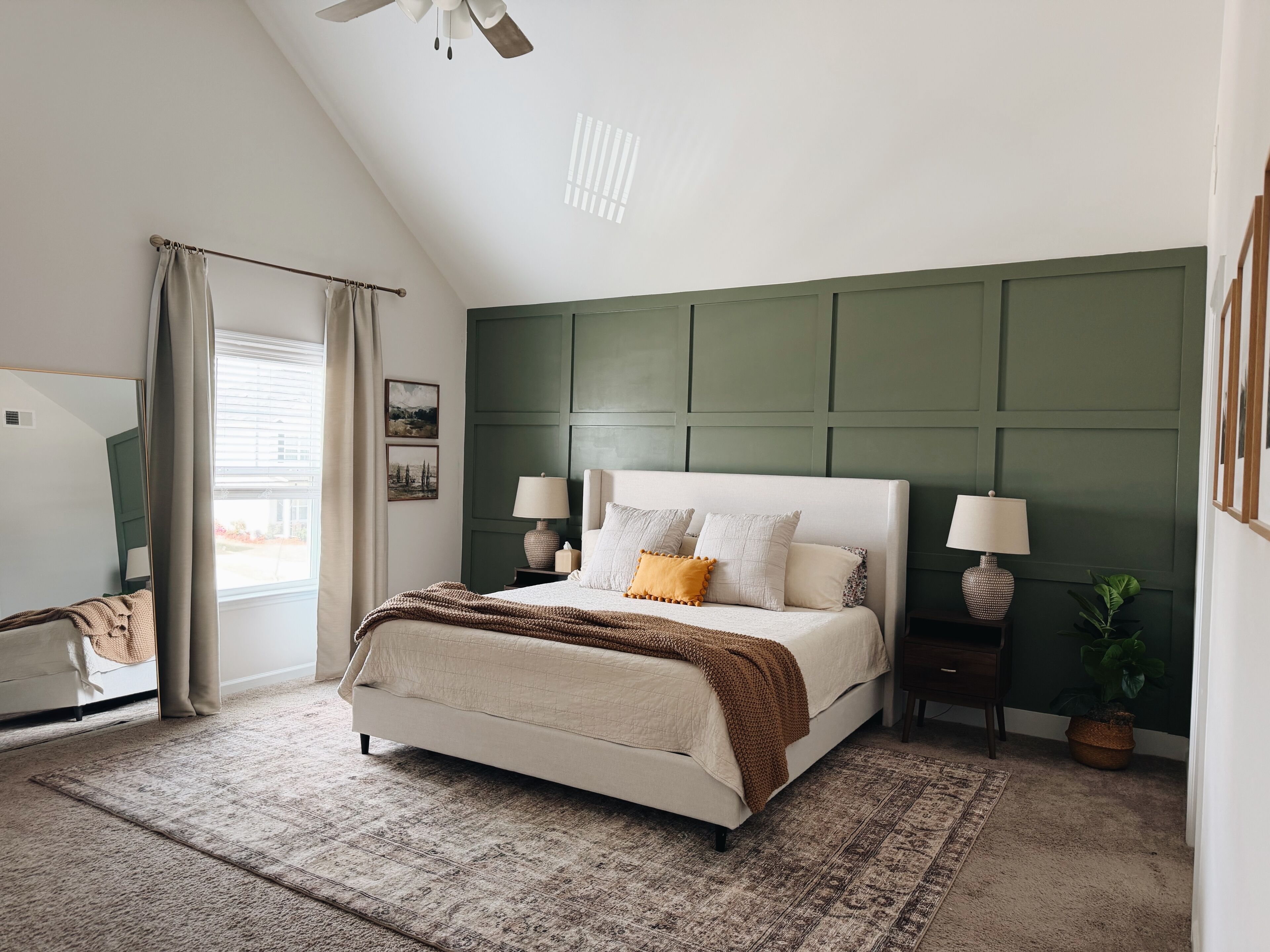 Une chambre avec un lit, deux tables de chevet avec des lampes, une grande fenêtre avec des rideaux, un ventilateur de plafond et un mur d’accent vert.