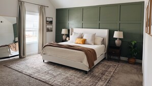 Une chambre avec un lit, deux tables de chevet avec des lampes, une grande fenêtre avec des rideaux, un ventilateur de plafond et un mur d’accent vert.
