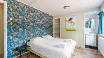 A bedroom with a floral wallpaper, a bed with white linens, a bedside table, a painting on the wall, and a bathroom with a sink and mirror.