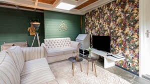 A living room with a green wall, a floral wallpaper, a tufted sofa, a television, and a small table with a vase. - Holiday Home in Gelderland Near Den Bramel (Lochem)
