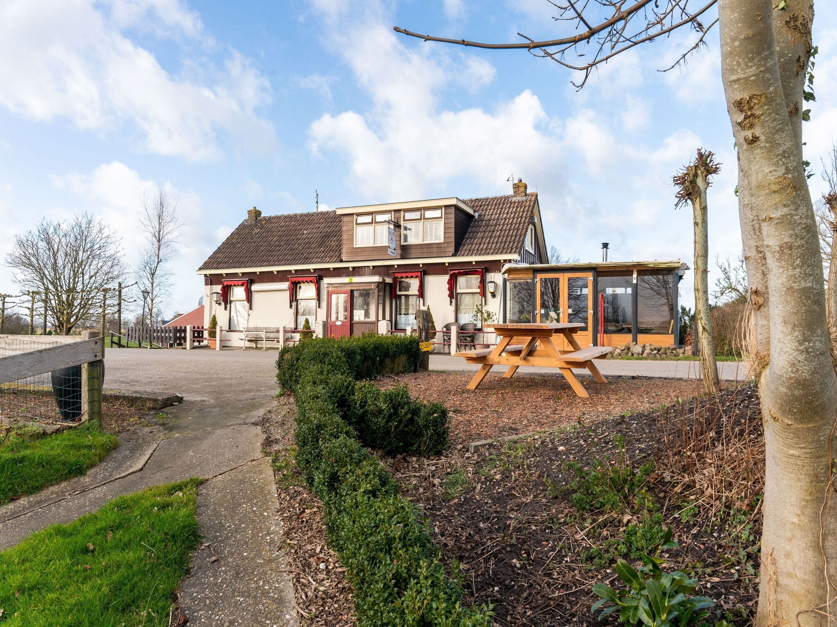 A two-story building with a traditional design, featuring a patio area with a wooden table and benches, surrounded by well-maintained hedges and a gravel path leading to the entrance.