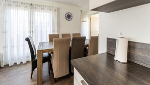 Un coin repas avec une table en bois et des chaises de couleur beige, une horloge au mur et une fenêtre ornée de rideaux légers.