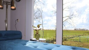 Una habitación moderna con un sofá azul, un florero verde con plantas y un ventanal con vistas a un campo verde.