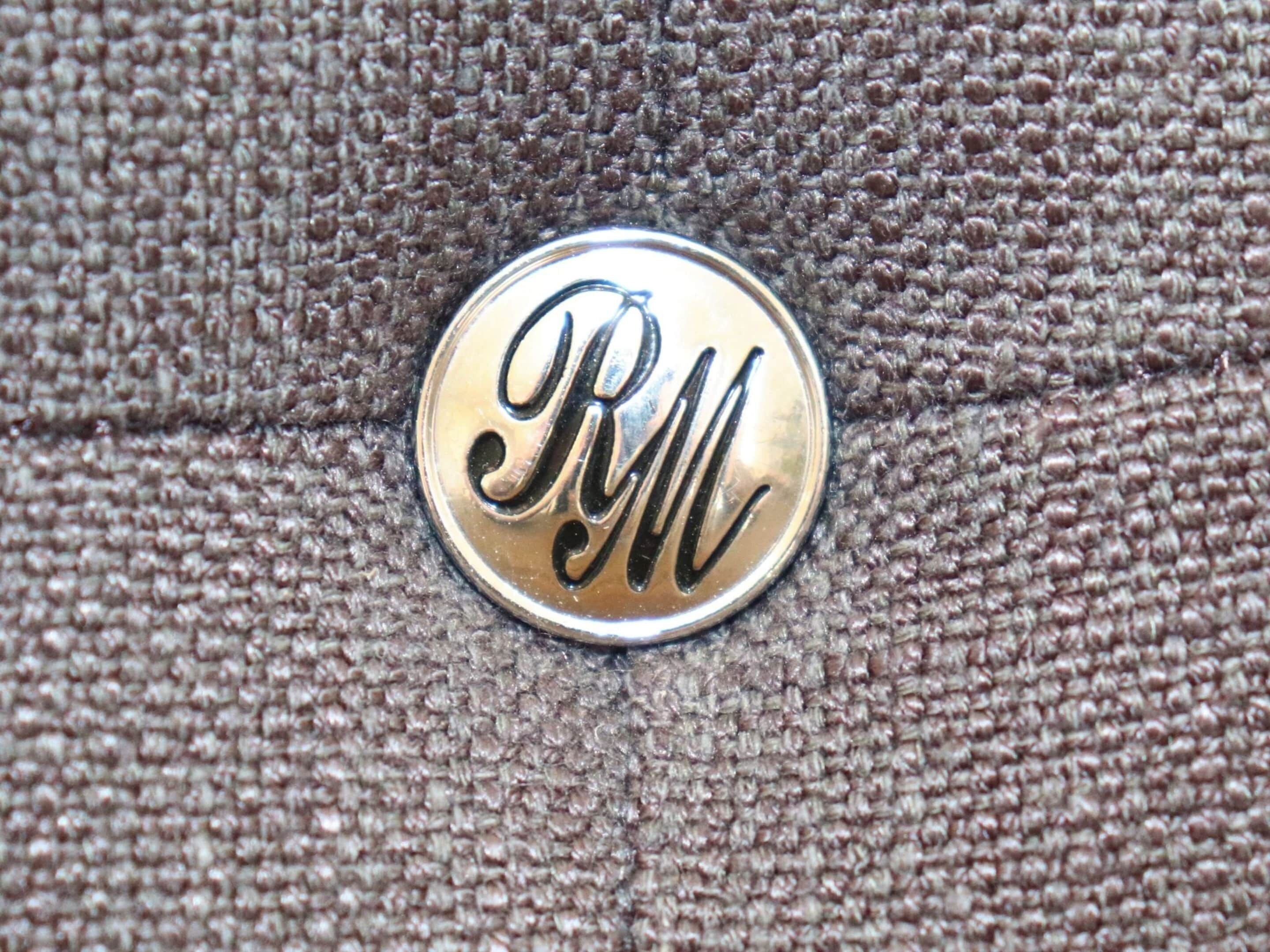 Ein metallischer Knopf mit den Buchstaben 'RM' auf einer stoffbespannten Oberfläche geprägt.