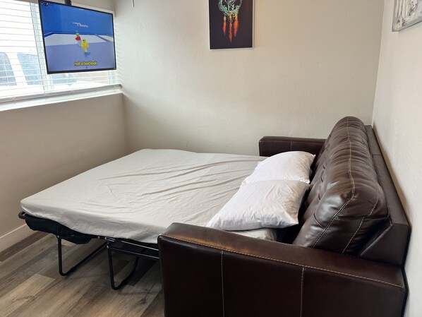 Une petite pièce avec un lit simple, un canapé en cuir marron, une fenêtre avec des stores et un téléviseur fixé au mur qui diffuse un match.