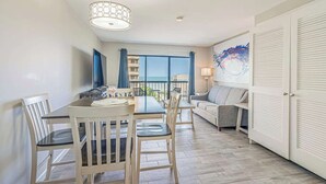 A modern hotel room with a dining area, a sofa, and a large window offering a view of the sea. - 1BR - Sleeps 6 - Steps to The Beach - Pool - 7500 (Myrtle Beach)