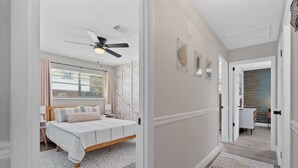 A bedroom with a bed, a ceiling fan, a window with curtains, and a hallway leading to another room. - POSH LIVING in Fort Walton Beach (Fort Walton Beach)