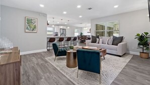 A modern living room with a white sofa, teal armchairs, and a wooden coffee table. A kitchen with bar stools is visible in the background. - POSH LIVING in Fort Walton Beach (Fort Walton Beach)