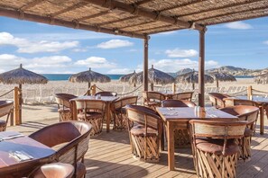 En restaurang vid stranden med träbord och stolar, parasoller med halmtak och utsikt över havet.