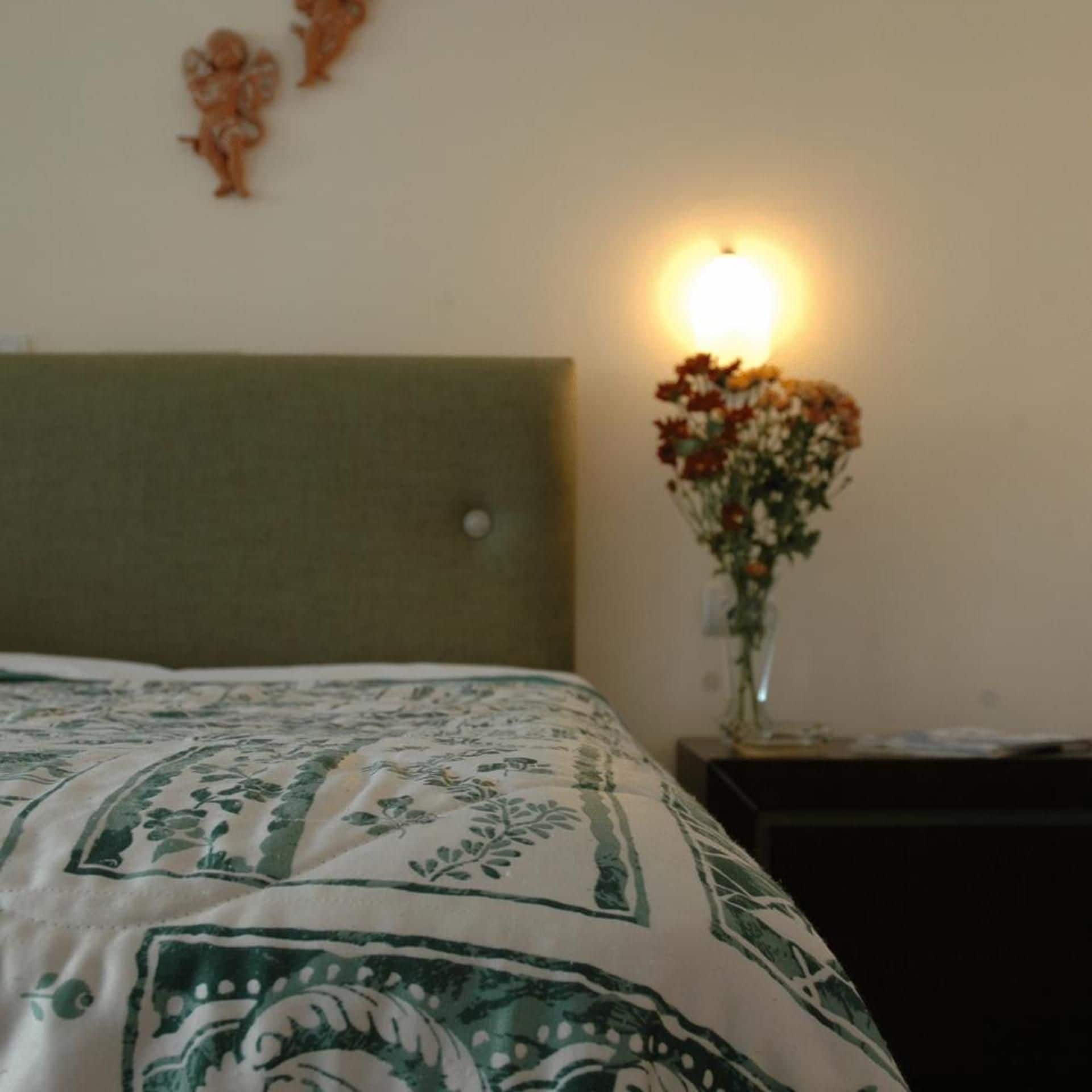 A bed with a patterned bedspread, a wall-mounted light, and a vase with flowers on a bedside table.