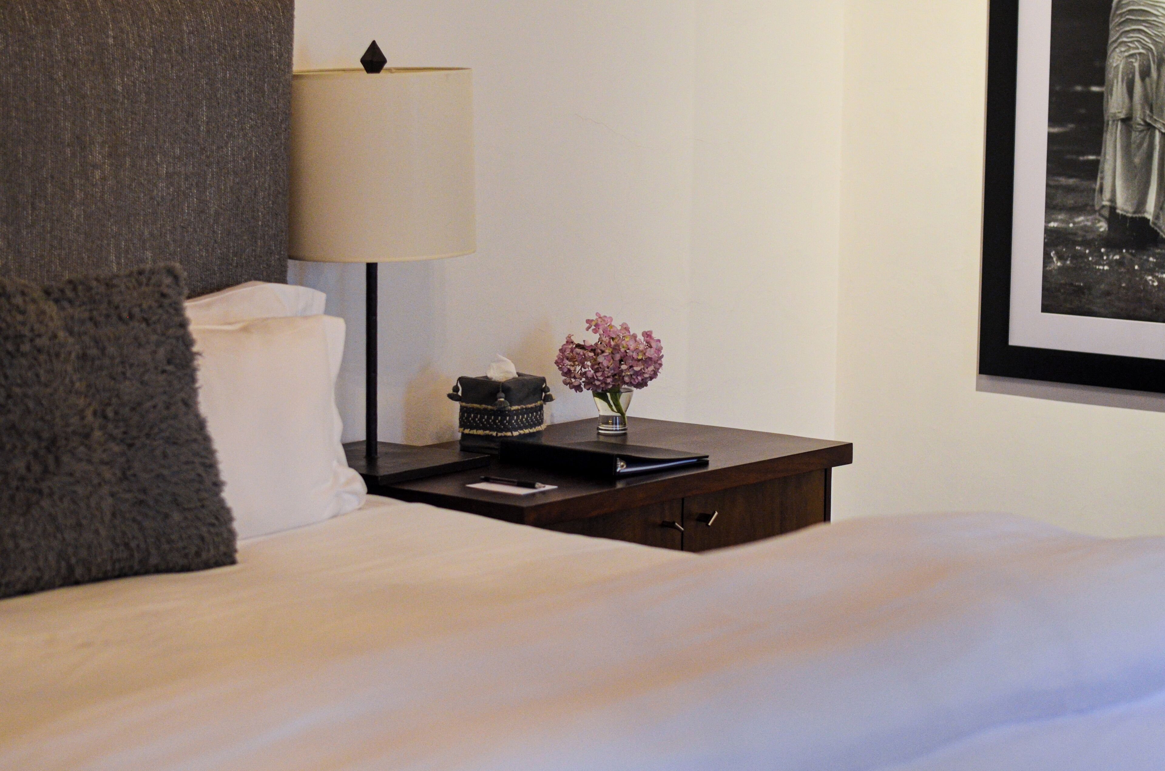 A neatly made bed with a pillow, a bedside table with a lamp, a vase with flowers, and a framed picture on the wall.
