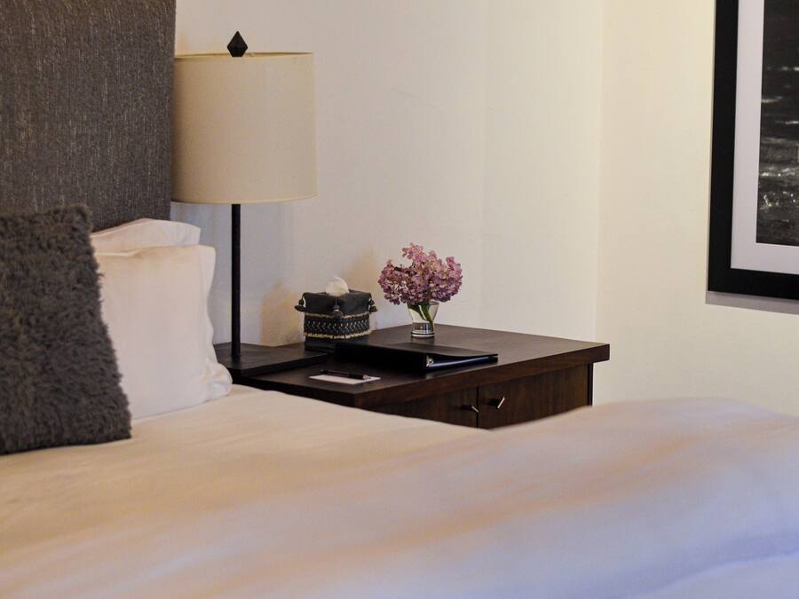 A neatly made bed with a pillow, a bedside table with a lamp, a vase with flowers, and a framed picture on the wall.