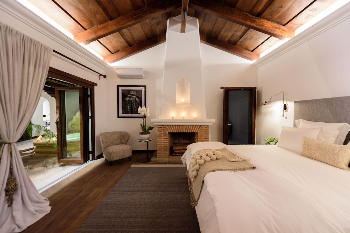A bedroom with a large bed, a fireplace, a ceiling fan, and a view of the outdoors.