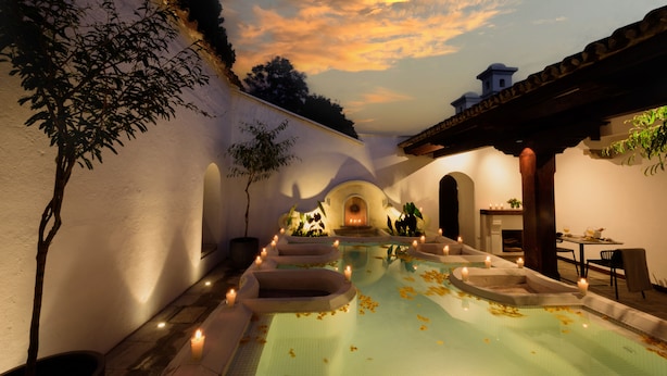 Un área de piscina tranquila con vista al atardecer, rodeada de paredes y arcos blancos, que incluye un comedor y plantas en macetas.
