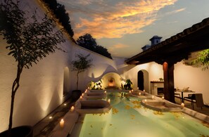 Un área de piscina tranquila con vista al atardecer, rodeada de paredes y arcos blancos, que incluye un comedor y plantas en macetas.