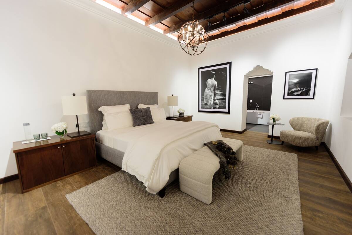 A bedroom with a large bed, bedside tables, a chandelier, framed pictures, and a chair.