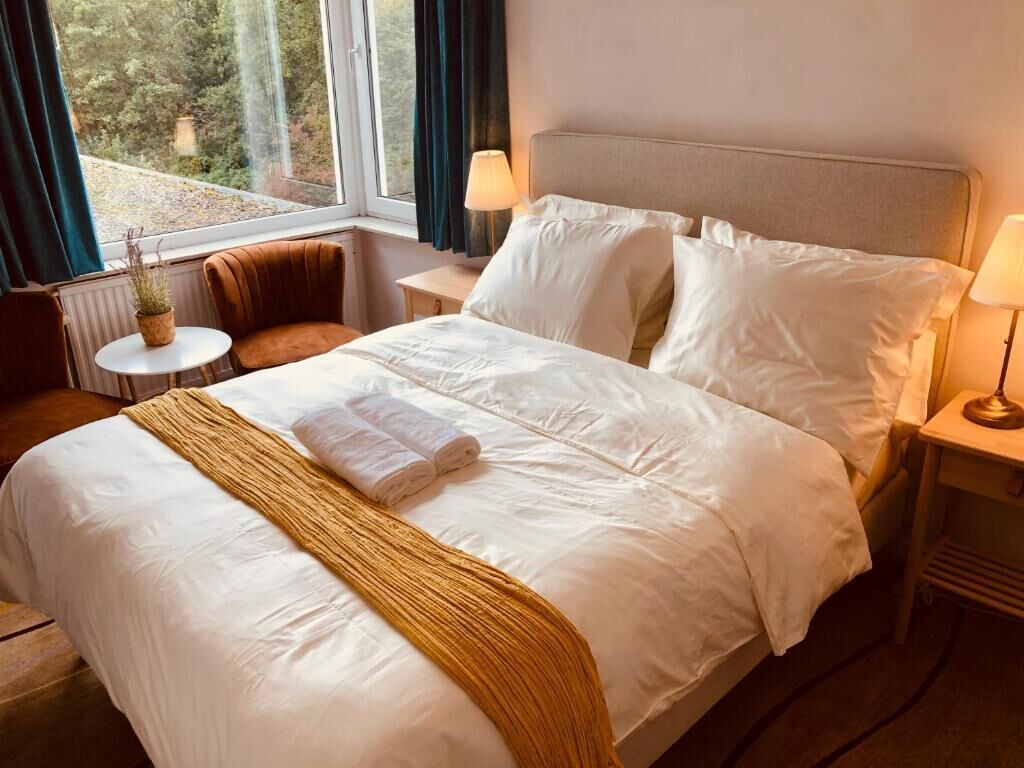 Ein ordentlich bezogenes Bett mit weißen Leinen und einer braunen Tagesdecke, ein kleiner runder Tisch mit einer Pflanze und ein Sessel in einem Zimmer mit Fenster und Nachttischlampe.