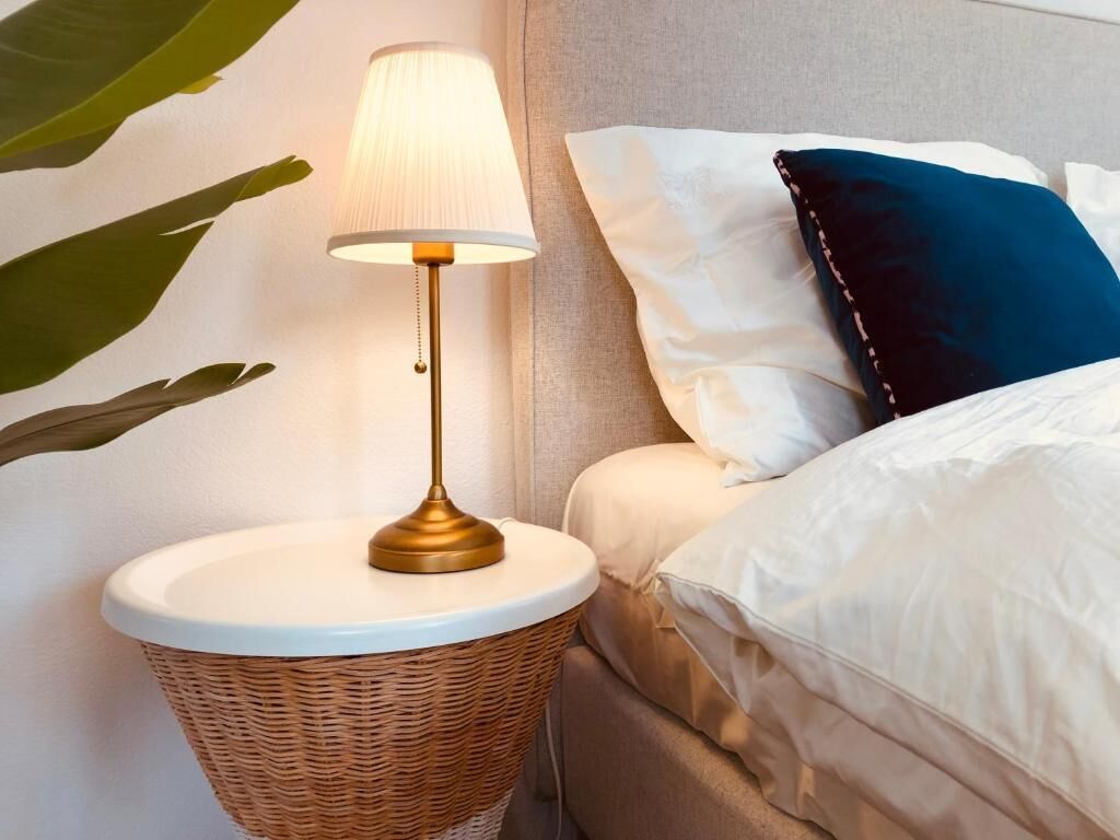 A bedside table with a lamp, a bed with white and blue pillows, and a wicker basket.
