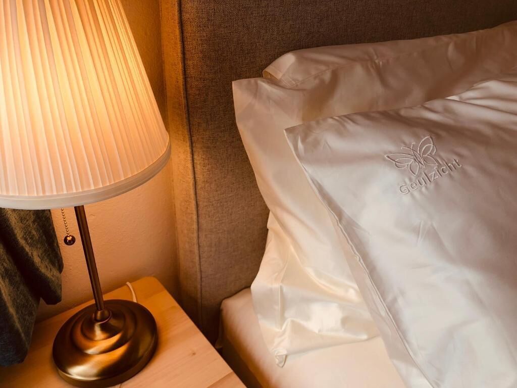 A neatly made bed with a white pillow featuring a logo, a bedside lamp on a wooden nightstand, and a grey headboard.