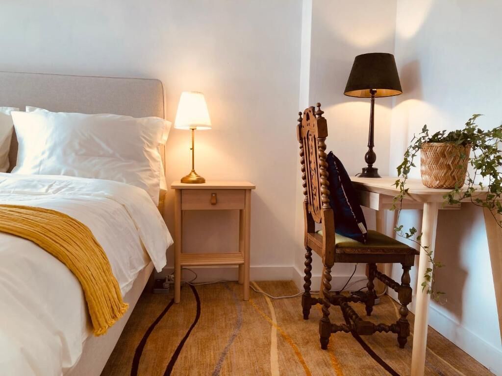A neatly arranged bedroom with a bed, bedside lamps, a wooden chair, and a small table with a plant.