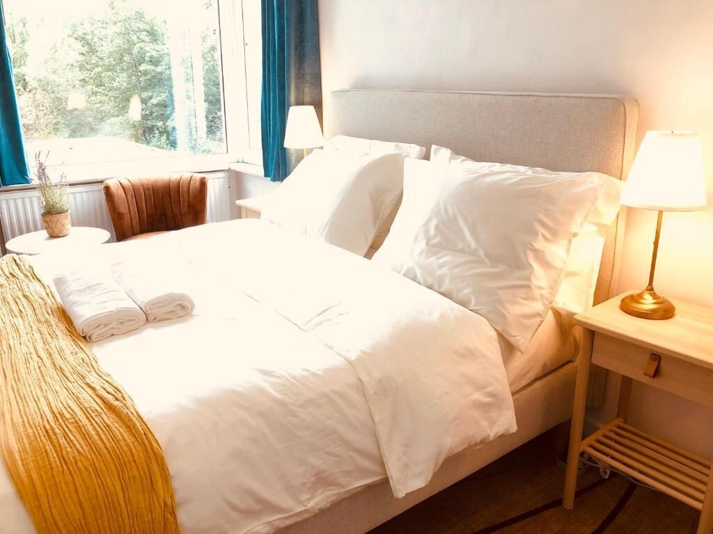 A neatly made bed with white linens, a bedside lamp, and a view of the outdoors through a window.