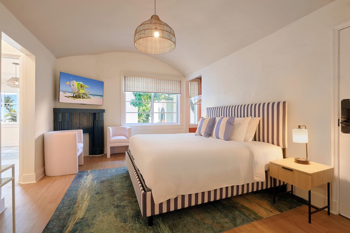 Un dormitorio con una cama grande, una mesita de noche con lámpara, una silla, un televisor en la pared y una ventana con persianas.