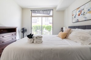A hotel room with a large bed, a dresser, a fan, and a window with a view of buildings and greenery.