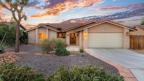 A single-story house with a garage, surrounded by a gravel driveway and landscaped with shrubs and a palm tree. - ✨Amazing Views✨Backyard Trails 🏜️Close to National Park, Shops, Restaurants ☕️ (Joshua Tree)