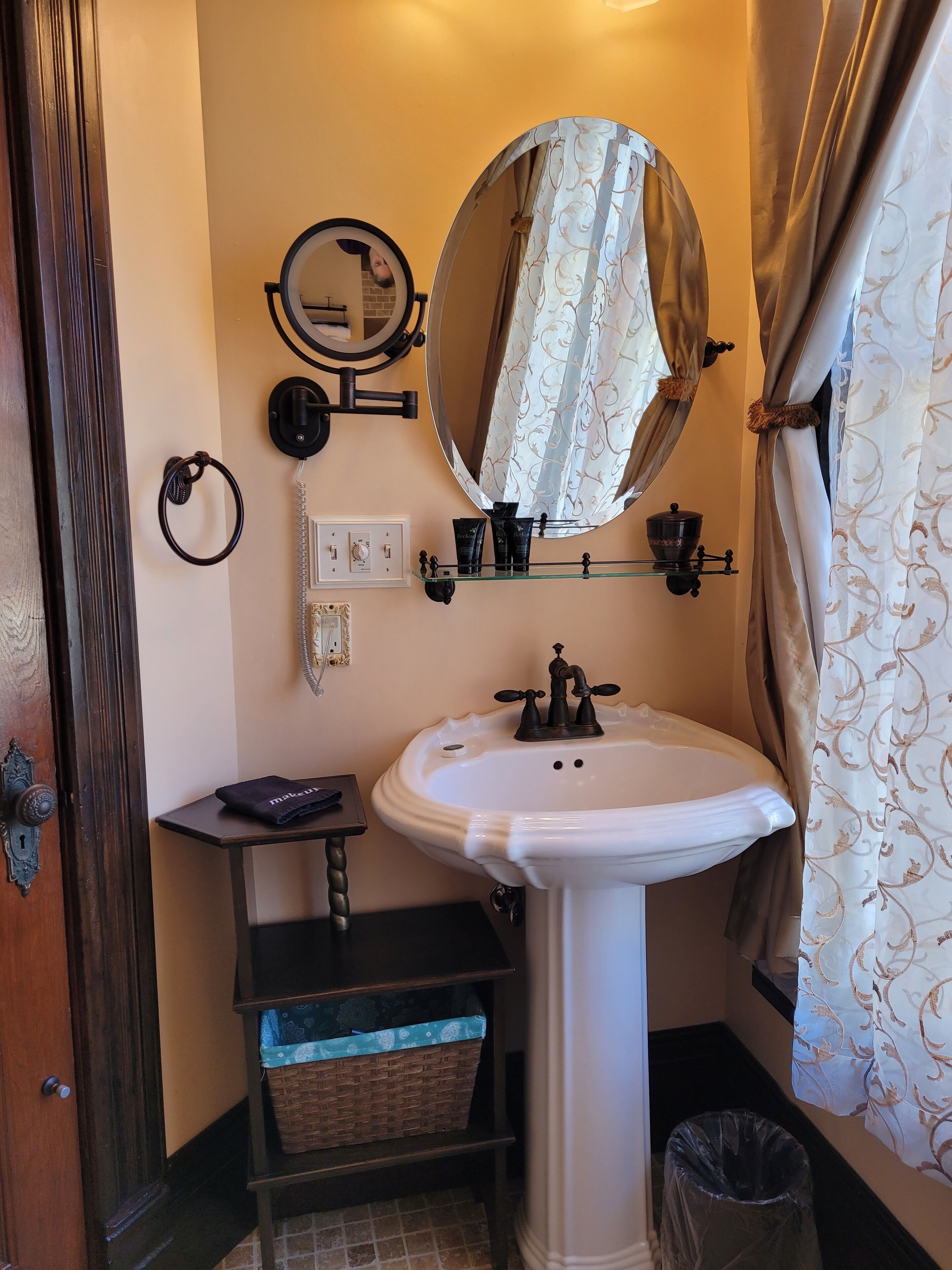 Un baño con lavamanos blanco, espejo, teléfono y repisas con artículos de tocador.