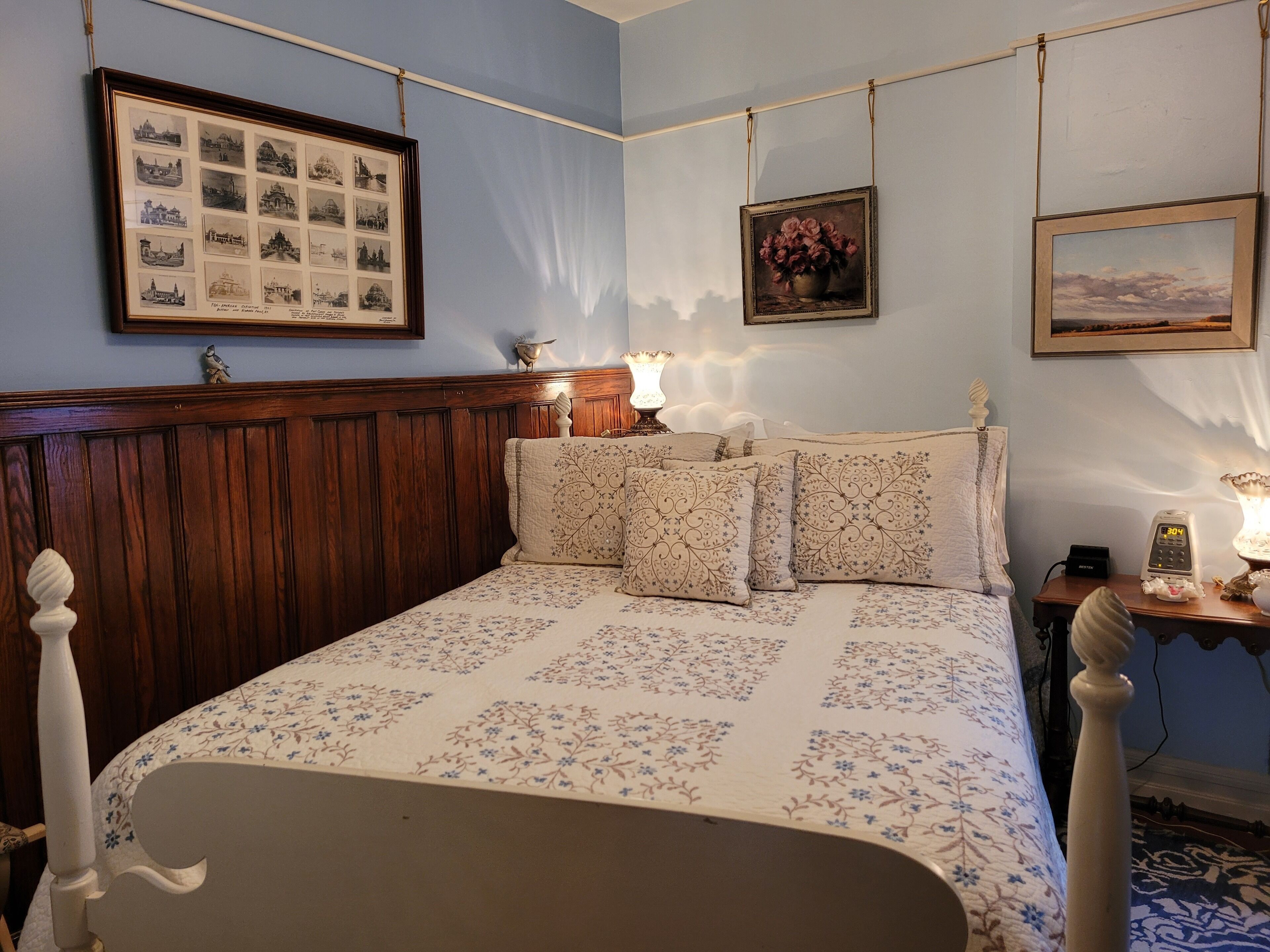 A bedroom with a wooden headboard, floral bedding, a bedside table with a lamp, and framed pictures on the wall.