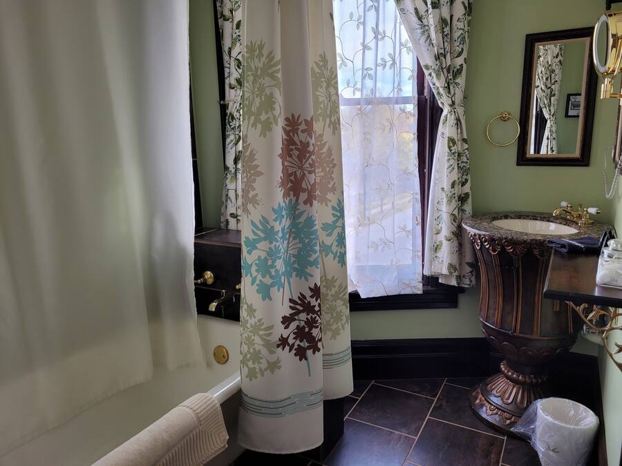 Un baño con un lavamanos antiguo, un espejo y una ventana con cortinas florales.