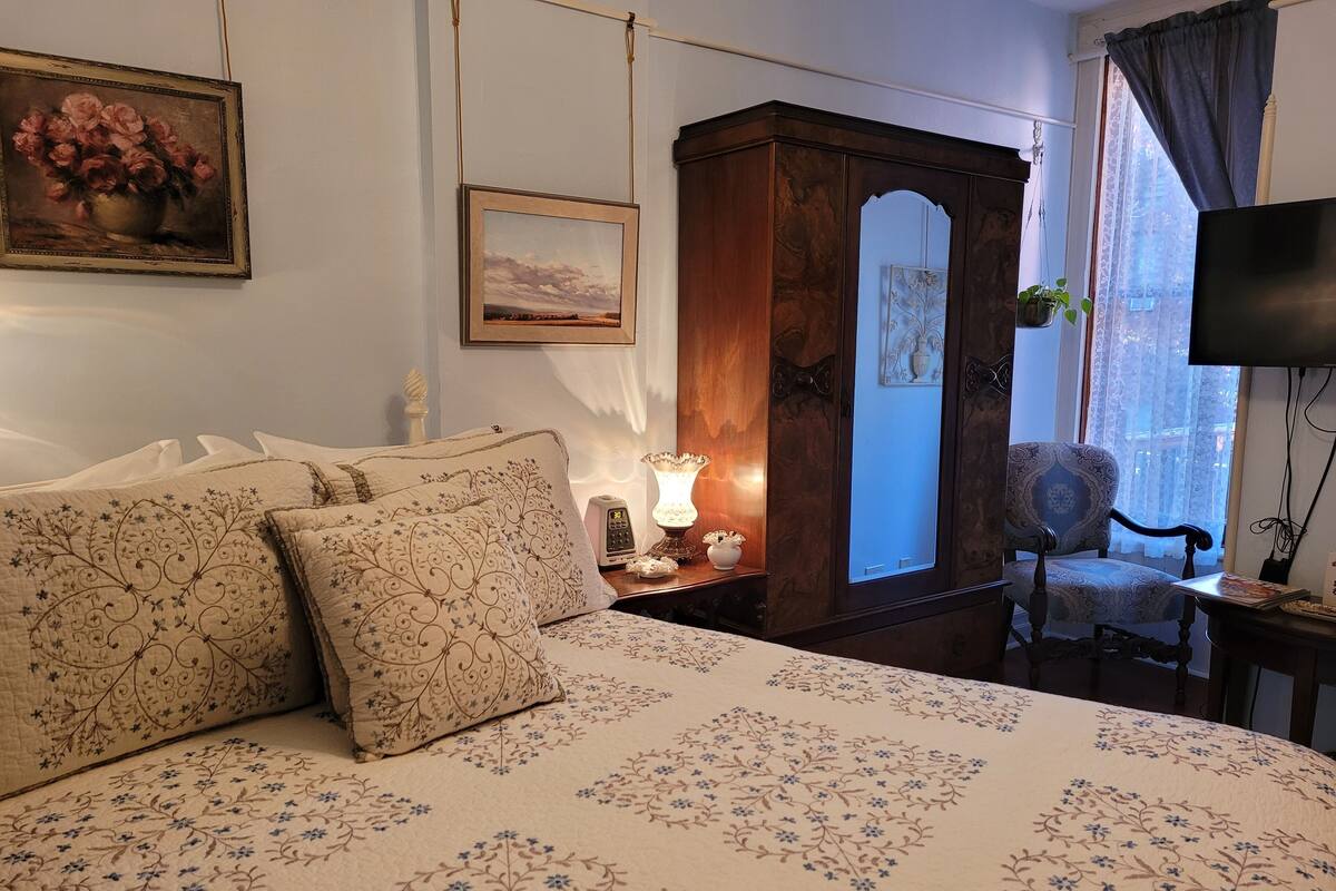 Quarto com uma cama grande, um guarda-roupa de madeira, uma cadeira, uma escrivaninha com uma luminária e uma televisão.