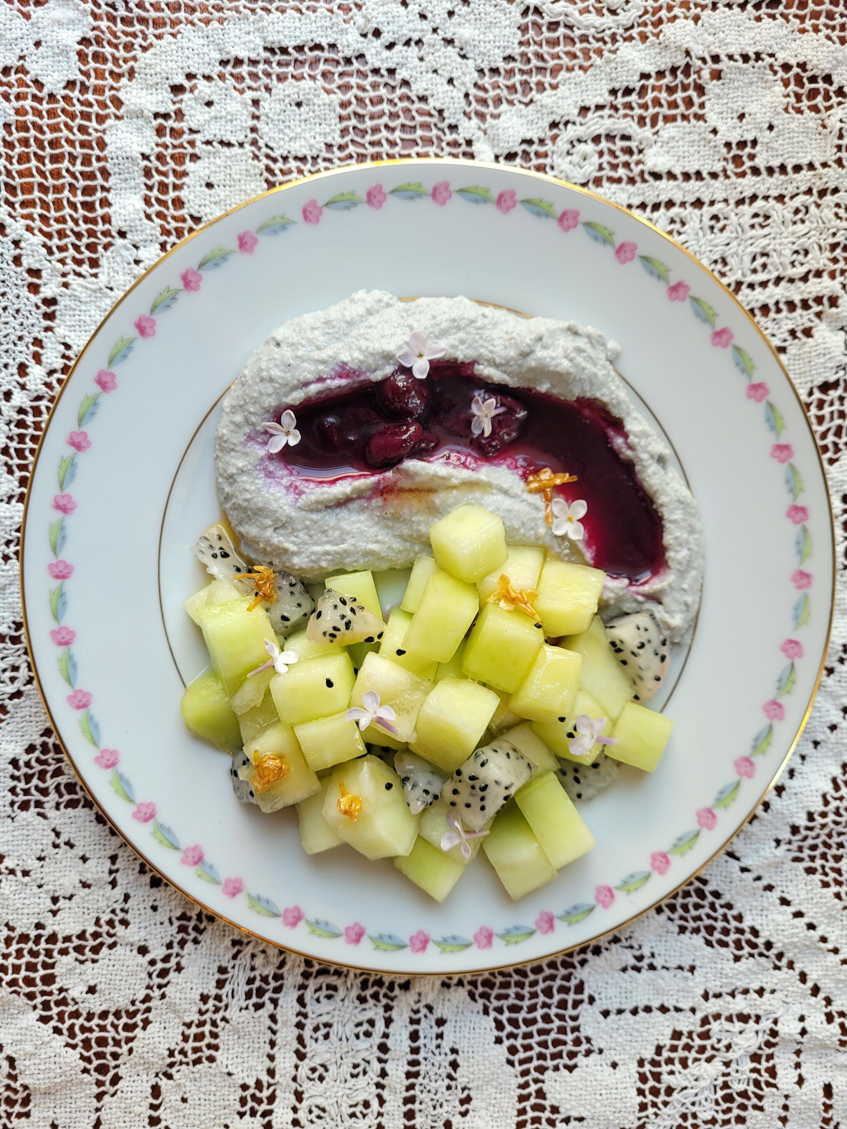 A plate of food with a creamy dessert, fruit cubes, and a decorative plate.
