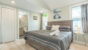 A bedroom with a large bed, a nightstand, a window with curtains, a painting on the wall, and a bathroom visible through an open door. - Private Waterfront Home Nestled In A Quiet Section Of Lake Hopatcong (Jefferson)