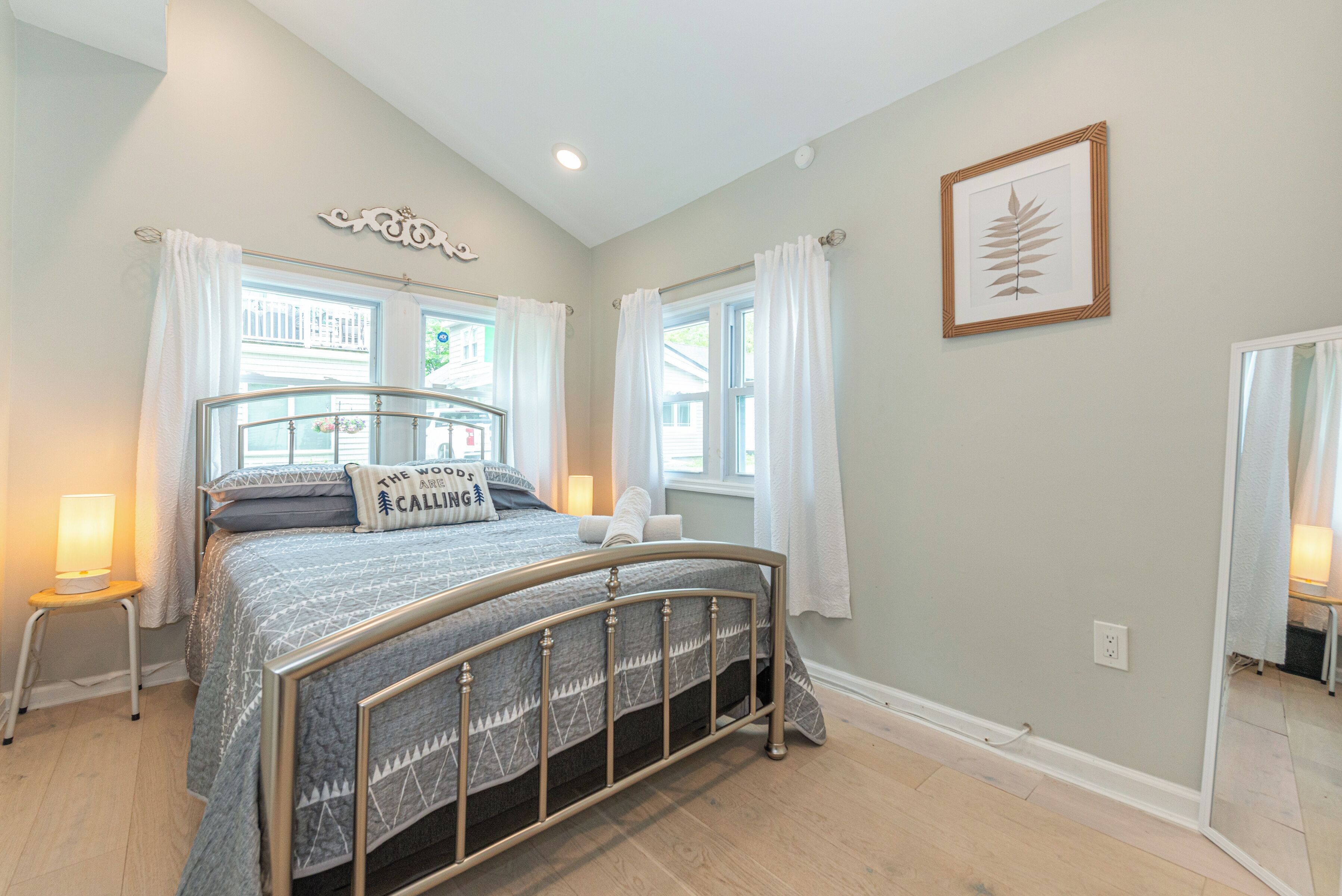 A bedroom with a bed, a bedside table, a window with curtains, a wall-mounted picture, and a mirror.