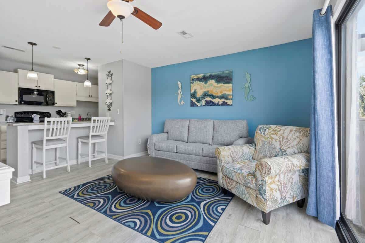 A modern living room with a grey sofa, patterned armchair, and ottoman. A kitchen with white cabinets and a microwave is visible in the background.