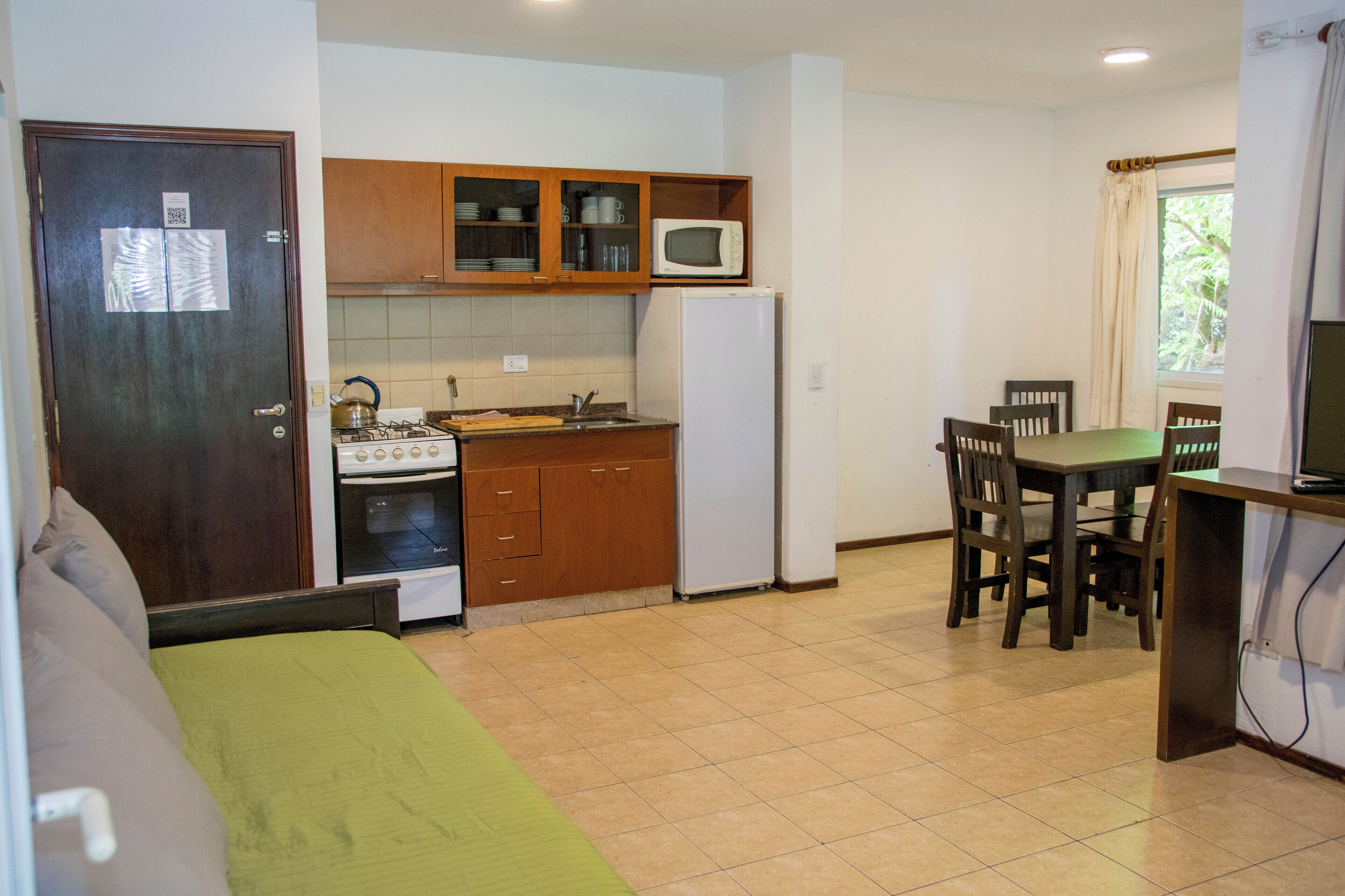 A compact kitchen with wooden cabinets, a microwave, a stove, and a refrigerator. There is a dining table with chairs and a green sofa in the living area.