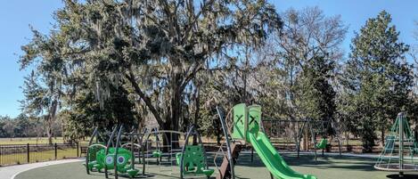 A playground with green equipment, including swings and a slide, surrounded by trees with Spanish moss.