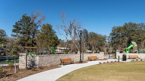 Un parque con un sendero pavimentado, bancos, un parque infantil con juegos verdes y un muro de ladrillo.