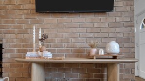 Un mur de briques sur lequel est fixé un téléviseur à écran plat, au-dessus d’une table en bois ornée de livres et d’objets décoratifs.